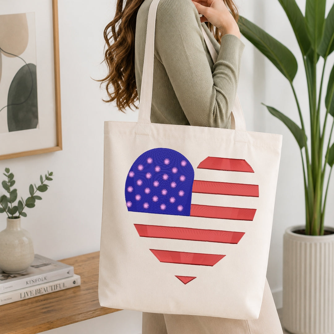 A woman wearing a sage green ribbed sweater carries a cream canvas tote bag with a heart-shaped American flag design featuring red and white stripes and a blue field with white stars. The background is a bright, modern room with a wooden table, books, a framed print, and a potted plant.