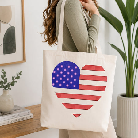 A woman wearing a sage green ribbed sweater carries a cream canvas tote bag with a heart-shaped American flag design featuring red and white stripes and a blue field with white stars. The background is a bright, modern room with a wooden table, books, a framed print, and a potted plant.