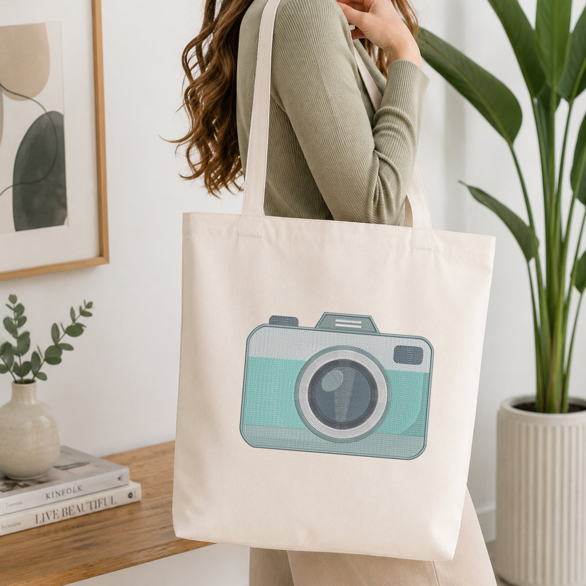 A woman wearing a sage green ribbed long-sleeve top carries a cream canvas tote bag with a printed illustration of a retro camera in mint green and gray. The scene is set in a bright, modern room with a wooden shelf, books, a small plant, and a large green potted plant in the background.