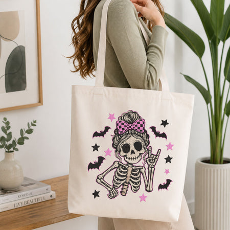 Woman carrying a white natural canvas tote bag featuring a cute skeleton messy bun machine embroidery design in a bright, modern interior setting