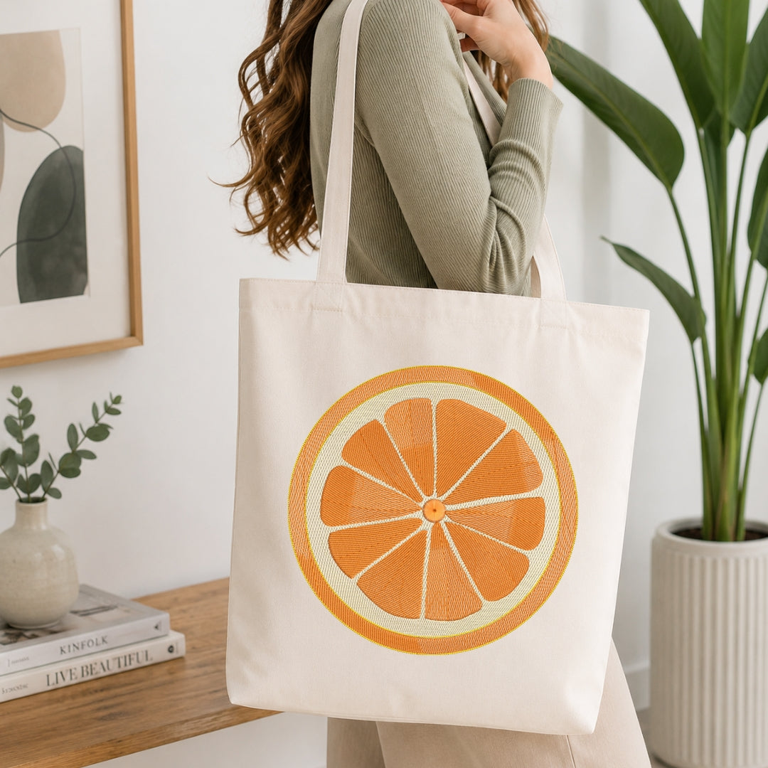 A person wearing a ribbed sage green top carries a cream canvas tote bag with a large orange slice graphic printed on the front, set in a bright, modern room with a wooden table, framed art, and a potted plant.