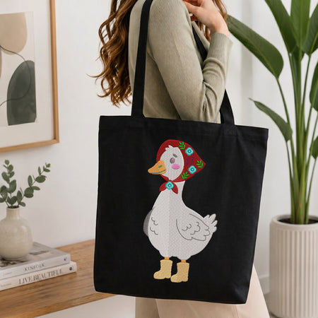 Person holding a black canvas tote bag featuring an embroidered cute goose wearing a red kerchief and boots.