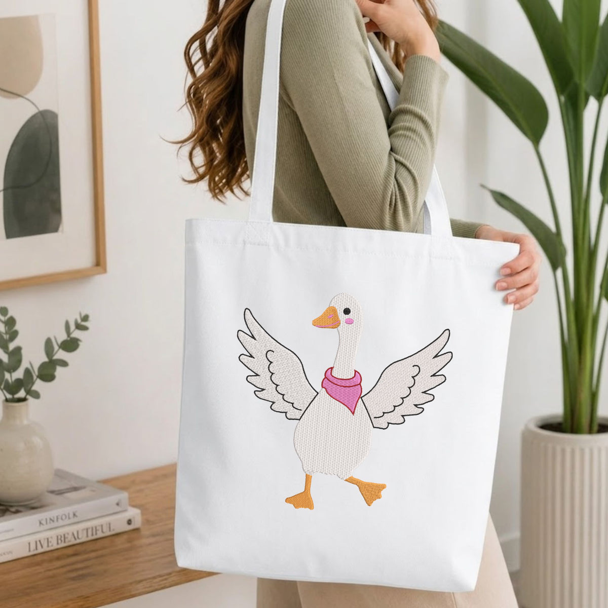 Woman carrying a white natural canvas tote bag featuring an embroidered dancing goose design in a bright, modern interior setting  .