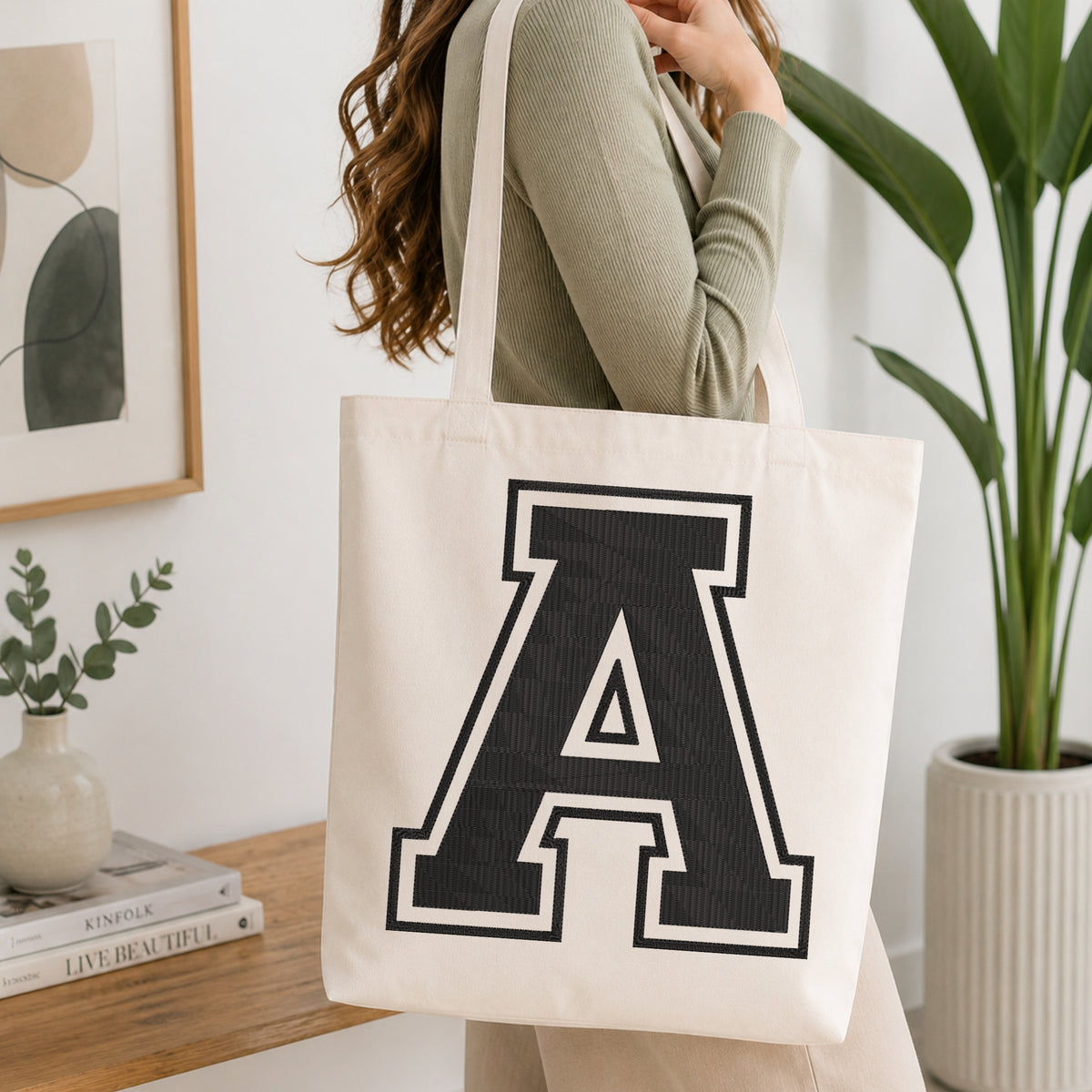 A woman wearing a sage green ribbed long-sleeve top carries a cream canvas tote bag with a large black collegiate letter 'A' printed on the front, set in a bright, modern room with a wooden console table and a potted green plant.