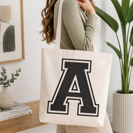A woman wearing a sage green ribbed long-sleeve top carries a cream canvas tote bag with a large black collegiate letter 'A' printed on the front, set in a bright, modern room with a wooden console table and a potted green plant.