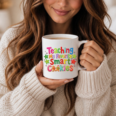 A woman in a cream knit sweater holds a white ceramic mug with colorful text that reads 'Teaching My Favorite Smart Cookies' decorated with small star and cookie illustrations.