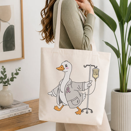 A person holding a natural canvas tote bag printed with an embroidered cute doctor duck illustration, set against a bright interior room background with green plants.