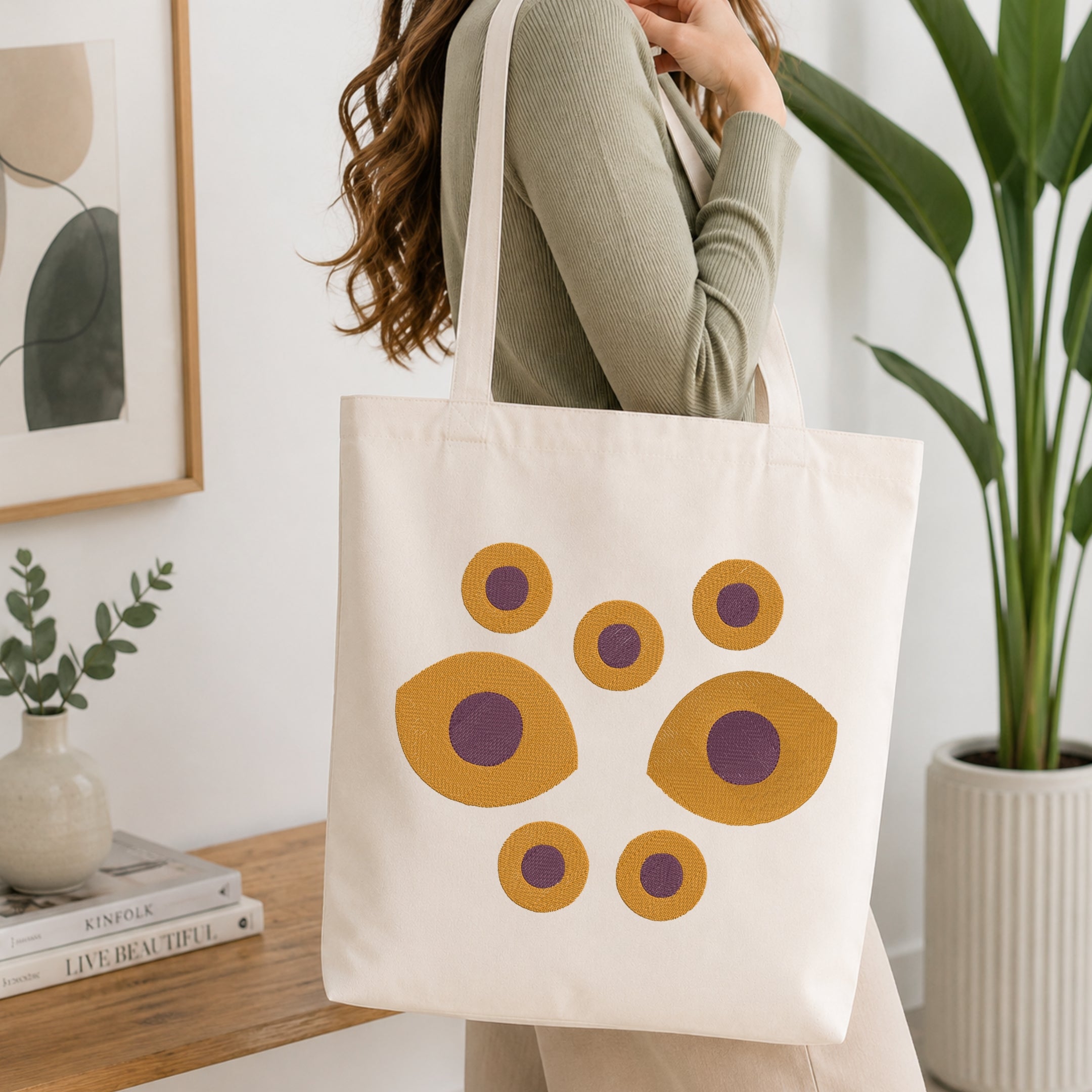 A woman wearing a sage green ribbed long-sleeve top carries a cream canvas tote bag decorated with a retro pattern of mustard yellow and purple circles, set in a bright, modern room with a wooden table, framed art, and a potted plant.