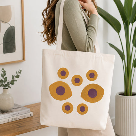 A woman wearing a sage green ribbed long-sleeve top carries a cream canvas tote bag decorated with a retro pattern of mustard yellow and purple circles, set in a bright, modern room with a wooden table, framed art, and a potted plant.