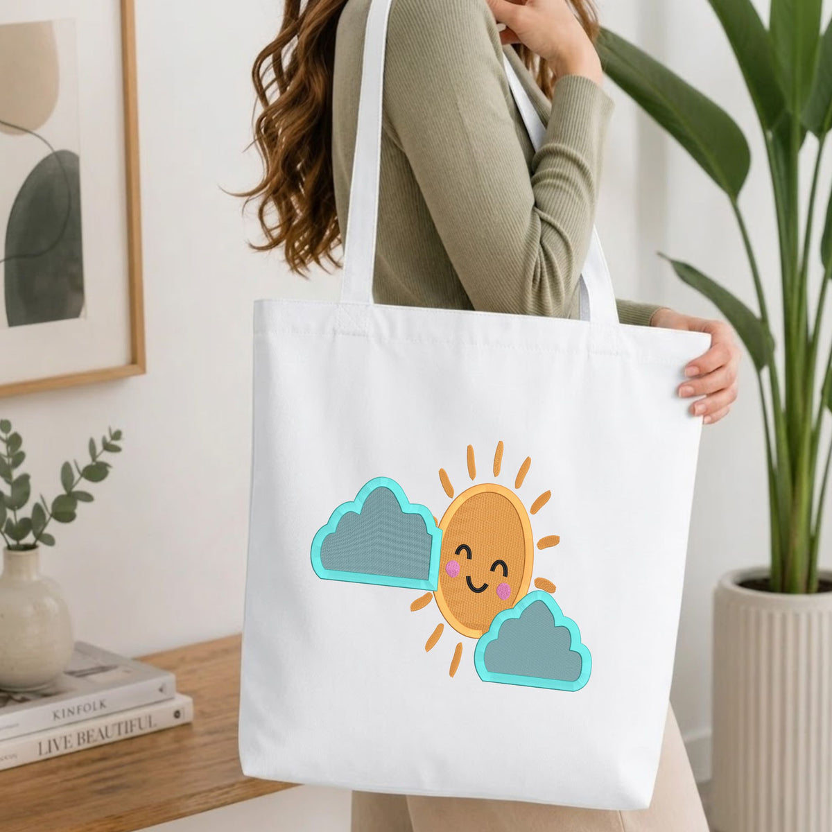 Woman carrying a white natural canvas tote bag featuring a cute smiling sun with blue clouds machine embroidery design in a bright, modern interior setting