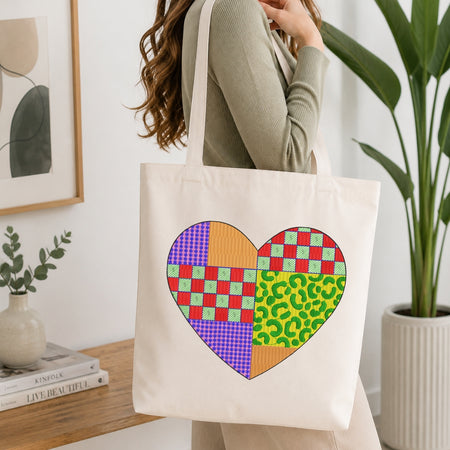 A person wearing a cream canvas tote bag displaying a heart motif composed of various colorful patchwork patterns.