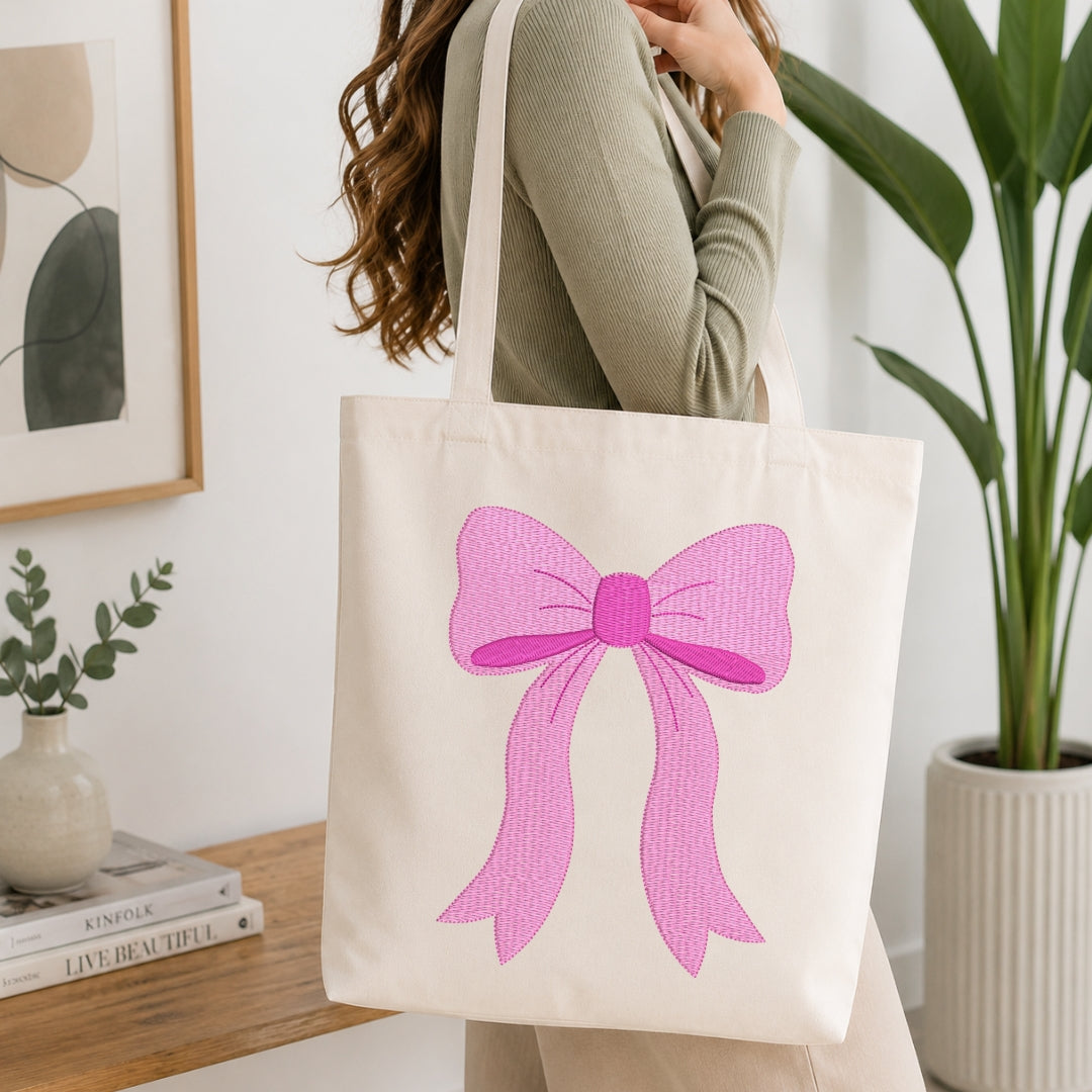 A woman wearing a sage green ribbed long-sleeve top carries a cream canvas tote bag with a large pink bow graphic on the front, set in a bright, modern room with a wooden table, framed art, and a potted plant.