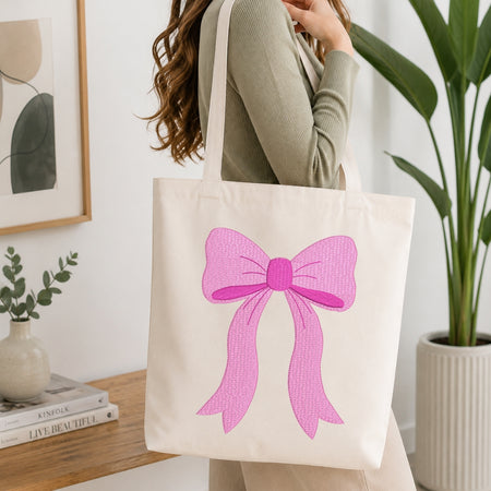 A woman wearing a sage green ribbed long-sleeve top carries a cream canvas tote bag with a large pink bow graphic on the front, set in a bright, modern room with a wooden table, framed art, and a potted plant.