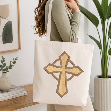 A person wearing a green top holds up a white canvas tote bag printed with the textured gold-yellow Celtic cross embroidery design, set against a bright, minimalist room background with indoor plants.