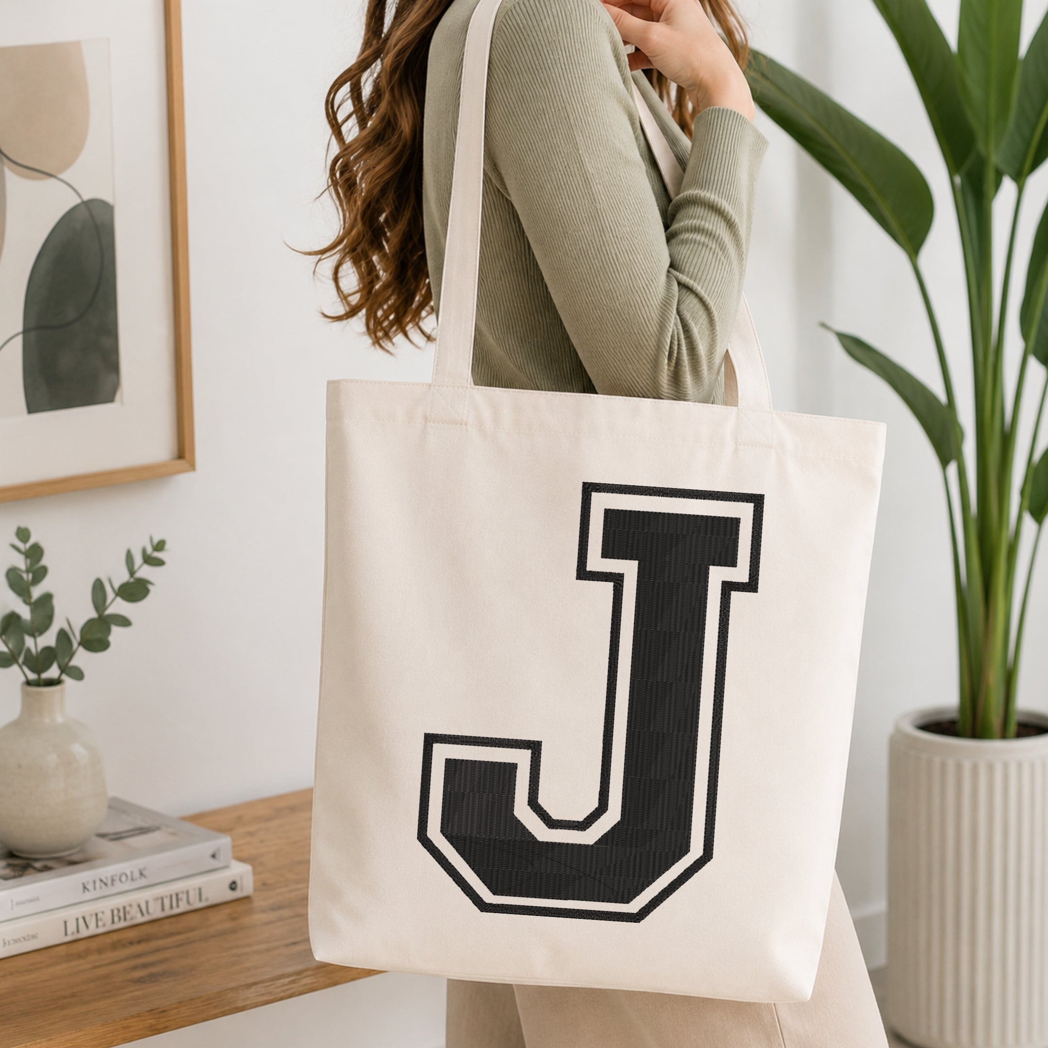 A woman wearing a sage green ribbed long-sleeve top carries a cream canvas tote bag with a large black collegiate letter 'J' outlined in white, set against a bright, modern interior background with a wooden console table, framed art, and a potted plant.