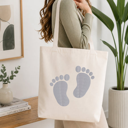 A woman wearing a sage green ribbed long-sleeve top carries a cream canvas tote bag with a light blue baby footprint graphic on the front, set in a bright, modern room with a wooden table, framed art, and a potted plant.