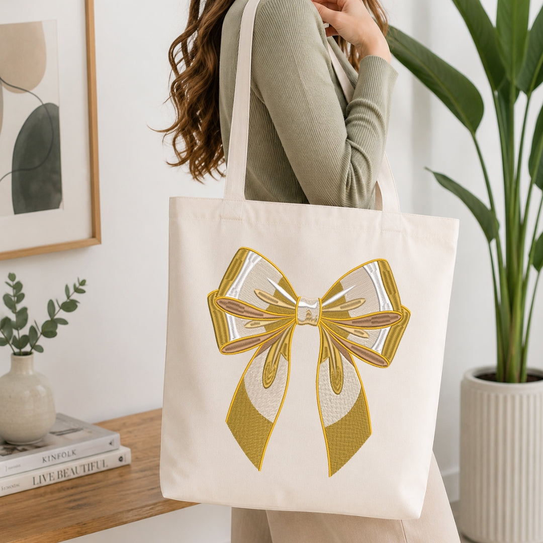 An off-white canvas tote bag with a large gold and white bow design, carried on the shoulder of a woman wearing a sage green ribbed sweater in a bright, modern room.