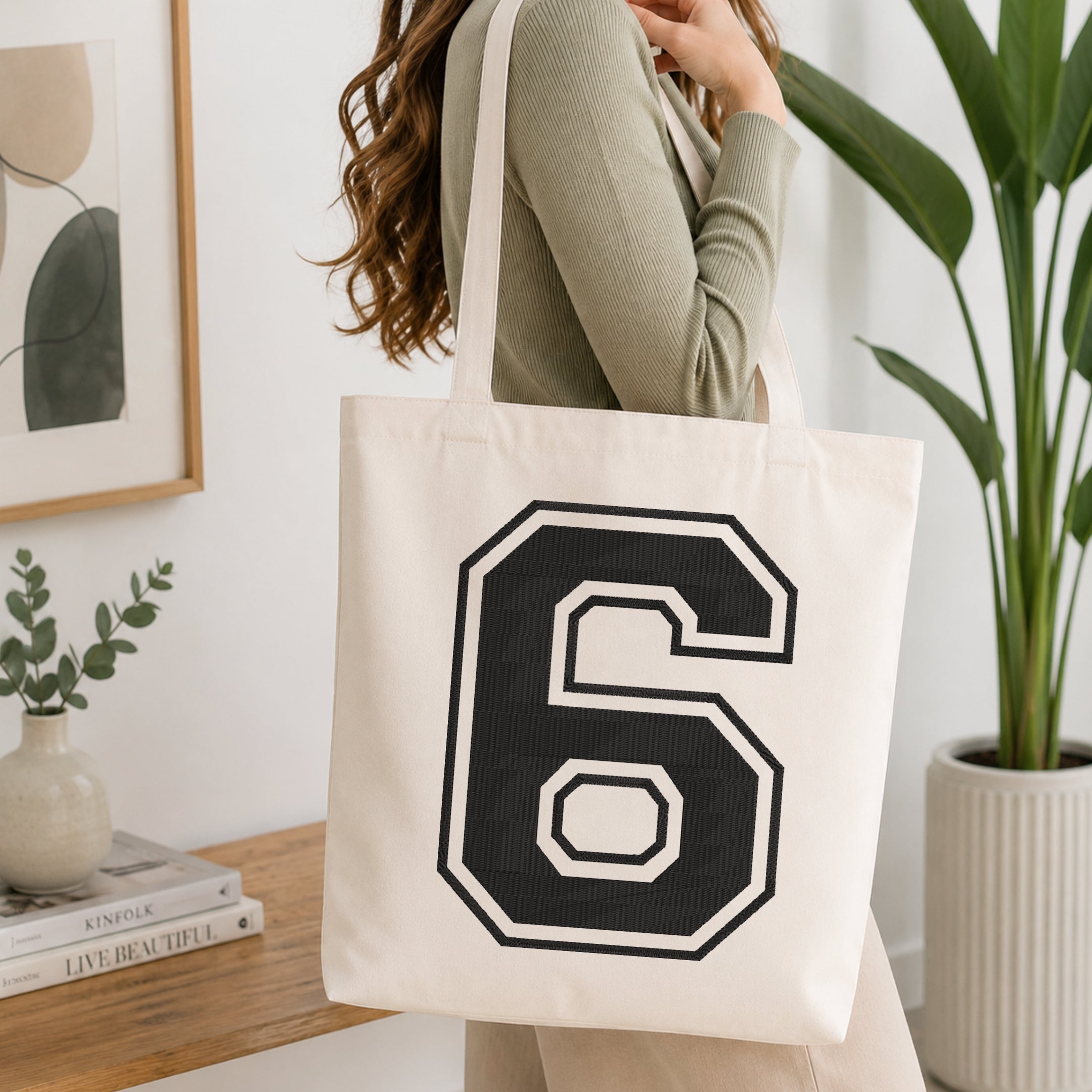 A woman wearing a sage green ribbed long-sleeve top carries a cream canvas tote bag with a large black and white number 6 printed on the front, set in a bright, modern room with a wooden console table and a potted plant.