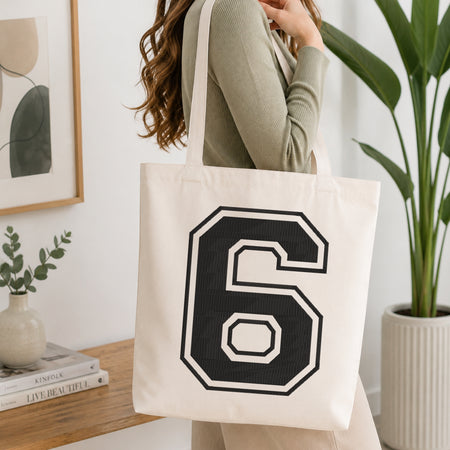 A woman wearing a sage green ribbed long-sleeve top carries a cream canvas tote bag with a large black and white number 6 printed on the front, set in a bright, modern room with a wooden console table and a potted plant.