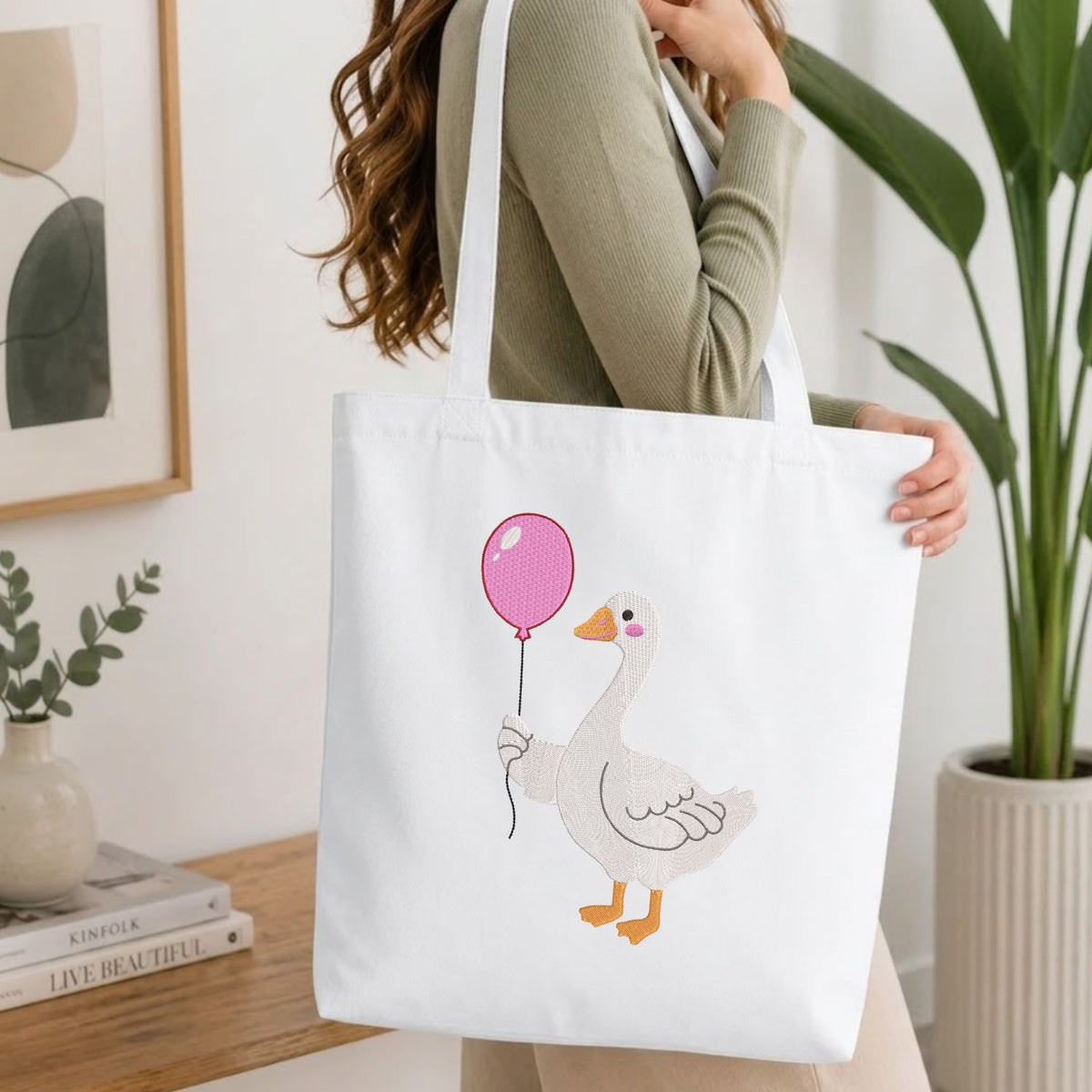 Woman carrying a white natural canvas tote bag featuring an embroidered cute duck in rain boots design in a bright, modern interior setting   