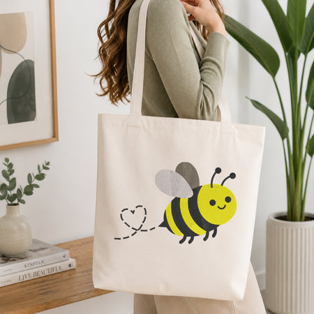 A woman wearing a sage green ribbed sweater carries a cream canvas tote bag with a cute yellow and black bumblebee graphic and a dashed heart tail. The scene is set in a bright, modern room with a wooden shelf, framed art, a small plant, and a large green potted plant.