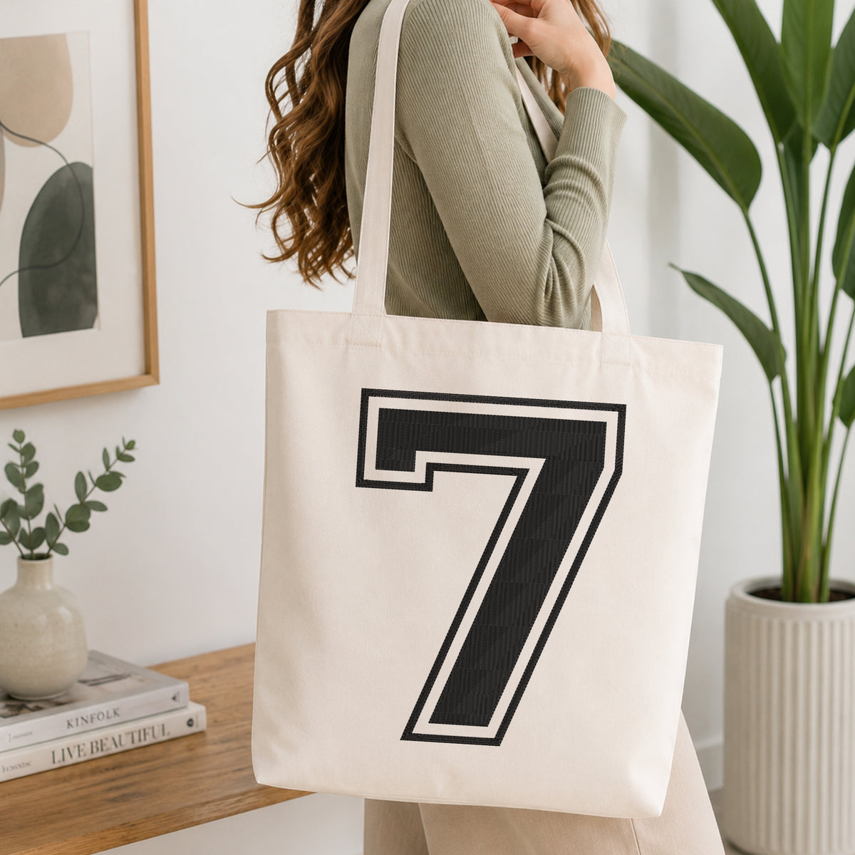 A woman wearing a sage green ribbed long-sleeve top carries a cream canvas tote bag with a large black and white number 7 printed on the front, set in a bright, modern room with a wooden console table and a potted plant.