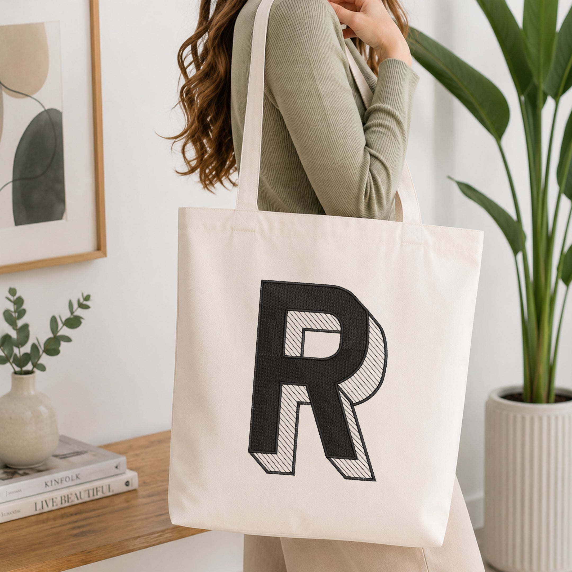 A woman wearing a sage green ribbed long-sleeve top carries a large off-white canvas tote bag with a bold black letter 'R' printed on the front, set in a bright, modern room with a wooden table, framed art, and a potted plant.