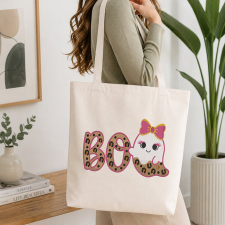 A person wearing a green long-sleeve top and carrying a natural canvas tote bag printed with the leopard print ghost "BOO" machine embroidery graphic.