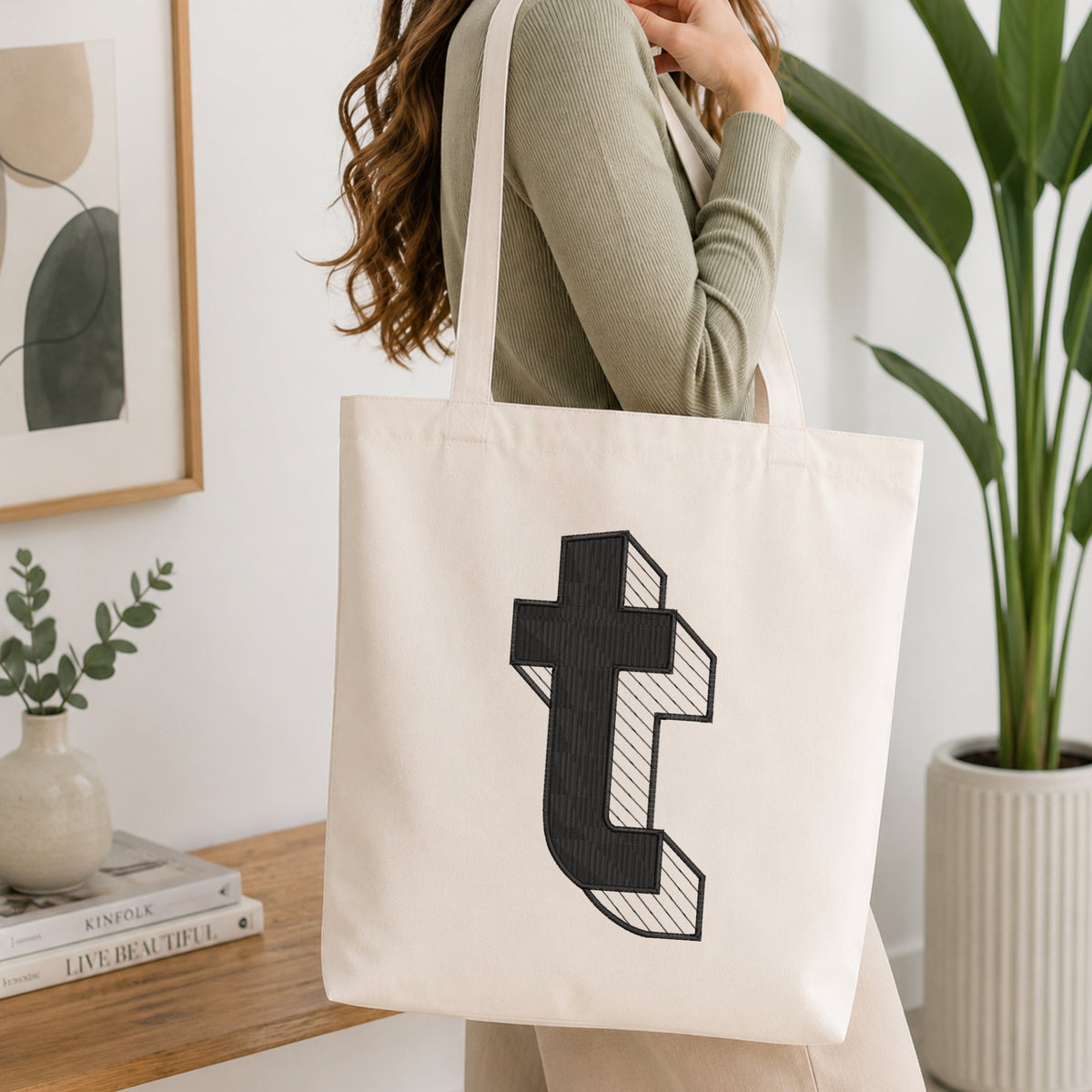 A woman wearing a sage green ribbed long-sleeve top carries a large off-white canvas tote bag with a black embroidered block letter 't' design on the front, set in a bright, modern room with a wooden table, books, and a potted plant.
