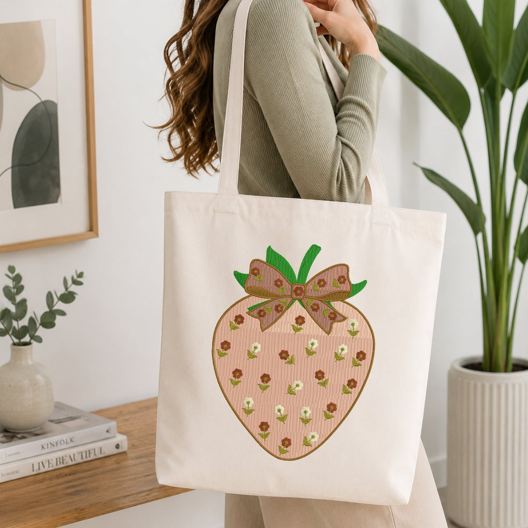 A woman wearing a sage green ribbed sweater carries a cream canvas tote bag with a large embroidered pink strawberry design featuring green leaves and small white flowers.