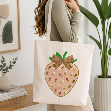 A woman wearing a sage green ribbed sweater carries a cream canvas tote bag with a large embroidered pink strawberry design featuring green leaves and small white flowers.