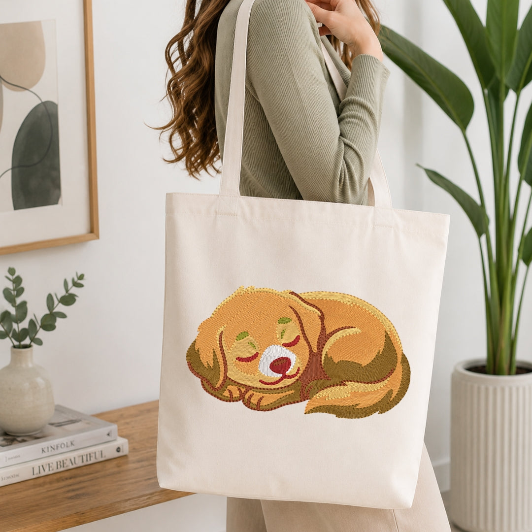 A woman wearing a sage green ribbed sweater carries a cream canvas tote bag with a printed illustration of a sleeping golden-brown dog. The scene is set in a bright, modern room with a wooden table, books, a small vase, and a large potted plant.