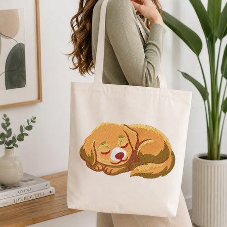 A woman wearing a sage green ribbed sweater carries a cream canvas tote bag with a printed illustration of a sleeping golden-brown dog. The scene is set in a bright, modern room with a wooden table, books, a small vase, and a large potted plant.