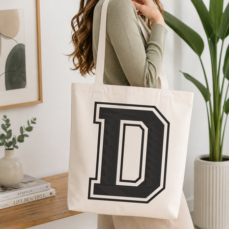 A person carrying a white canvas tote bag featuring a large embroidered collegiate-style black letter 'D' with a white border outline, set in a bright room with green house plants.