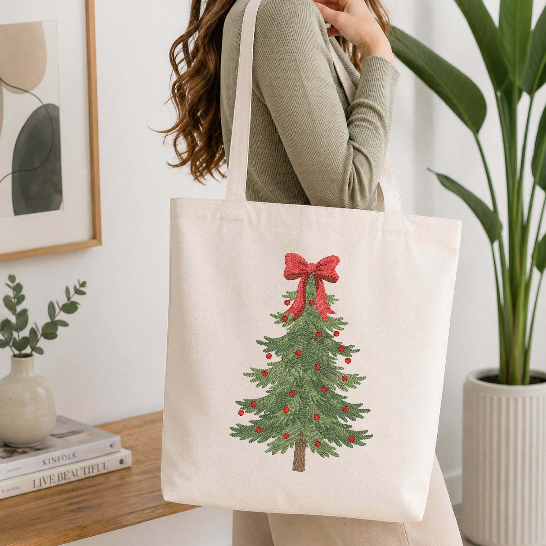 A woman wearing a sage green ribbed sweater carries a cream canvas tote bag with a printed Christmas tree design featuring a red bow and ornaments, set in a bright, modern room with a wooden table and potted plants.