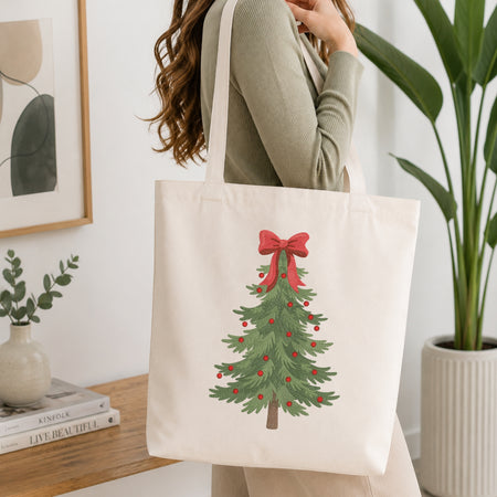 A woman wearing a sage green ribbed sweater carries a cream canvas tote bag with a printed Christmas tree design featuring a red bow and ornaments, set in a bright, modern room with a wooden table and potted plants.