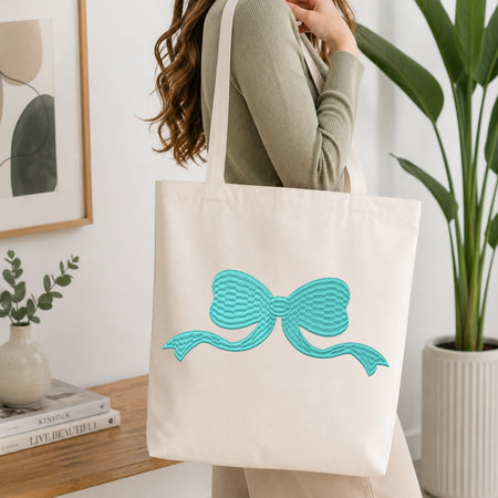 A person wearing a ribbed sage green top carries a large off-white canvas tote bag with a turquoise bow design on the front, set in a bright, modern room with a wooden table, books, and a potted plant.