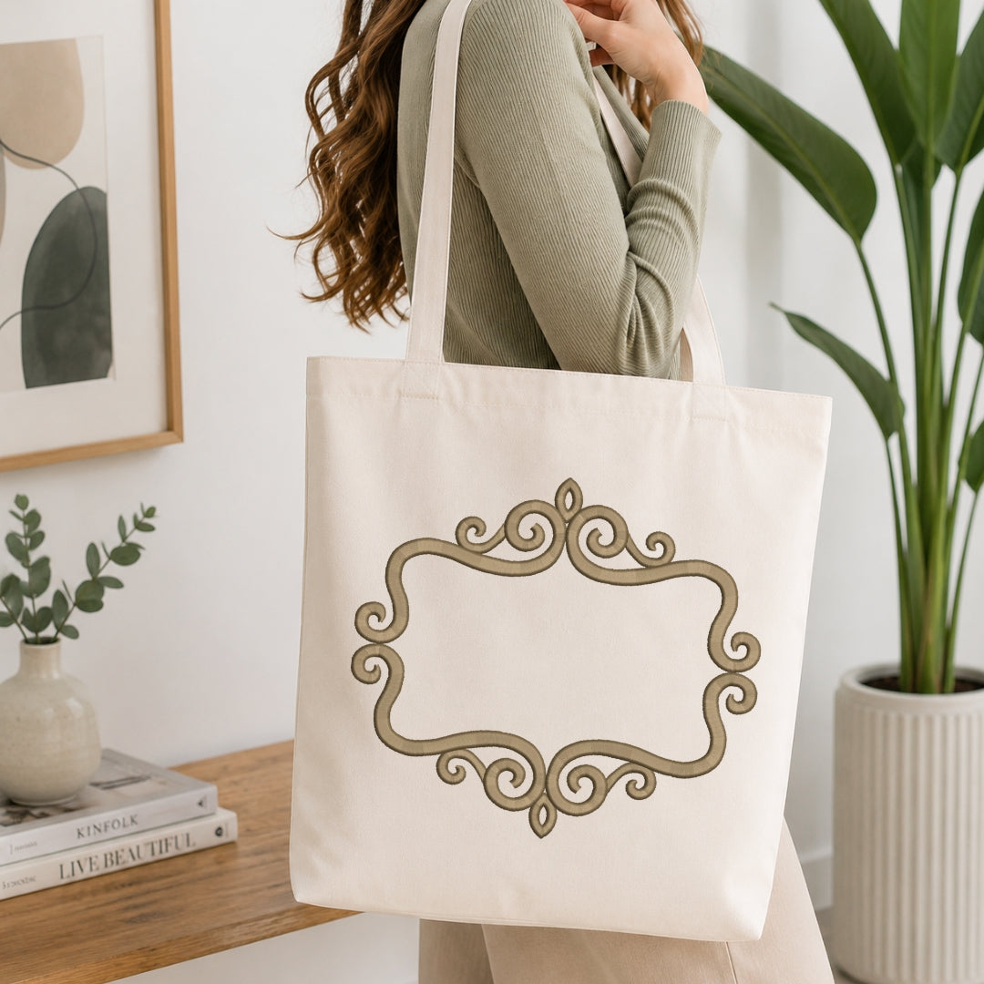 A woman wearing a sage green ribbed sweater carries a cream canvas tote bag with a large gold ornate frame design on the front, set in a bright, modern room with a wooden table, books, and a potted plant.