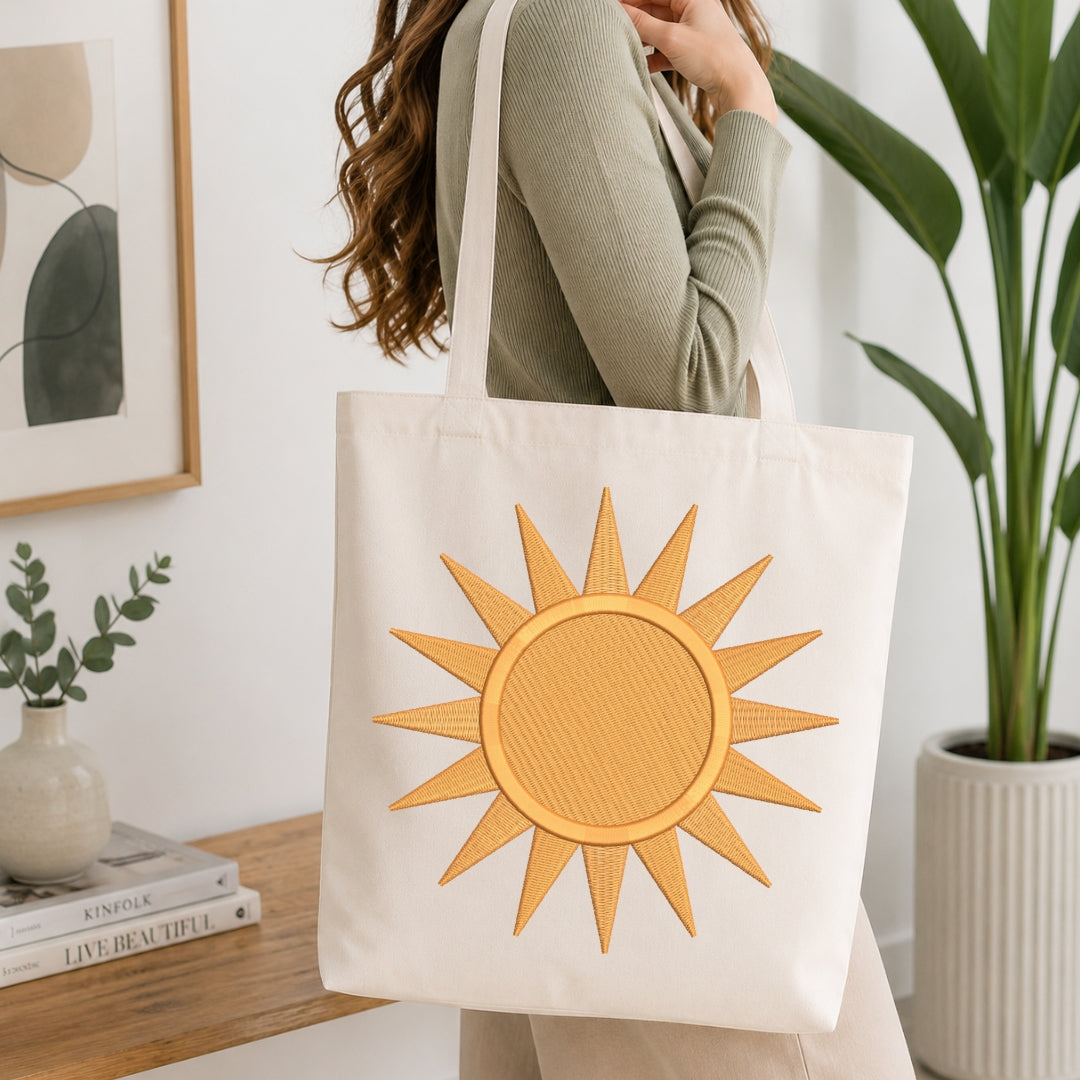 A woman wearing a sage green ribbed sweater carries a cream canvas tote bag with a large yellow-orange sun applique on the front, set in a bright, modern room with a wooden table, books, and a potted plant.