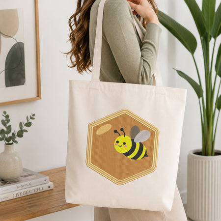 A woman wearing a sage green ribbed long-sleeve top carries a cream canvas tote bag with a cartoon bee design on the front, set in a bright, modern room with a wooden table, books, and a potted plant.