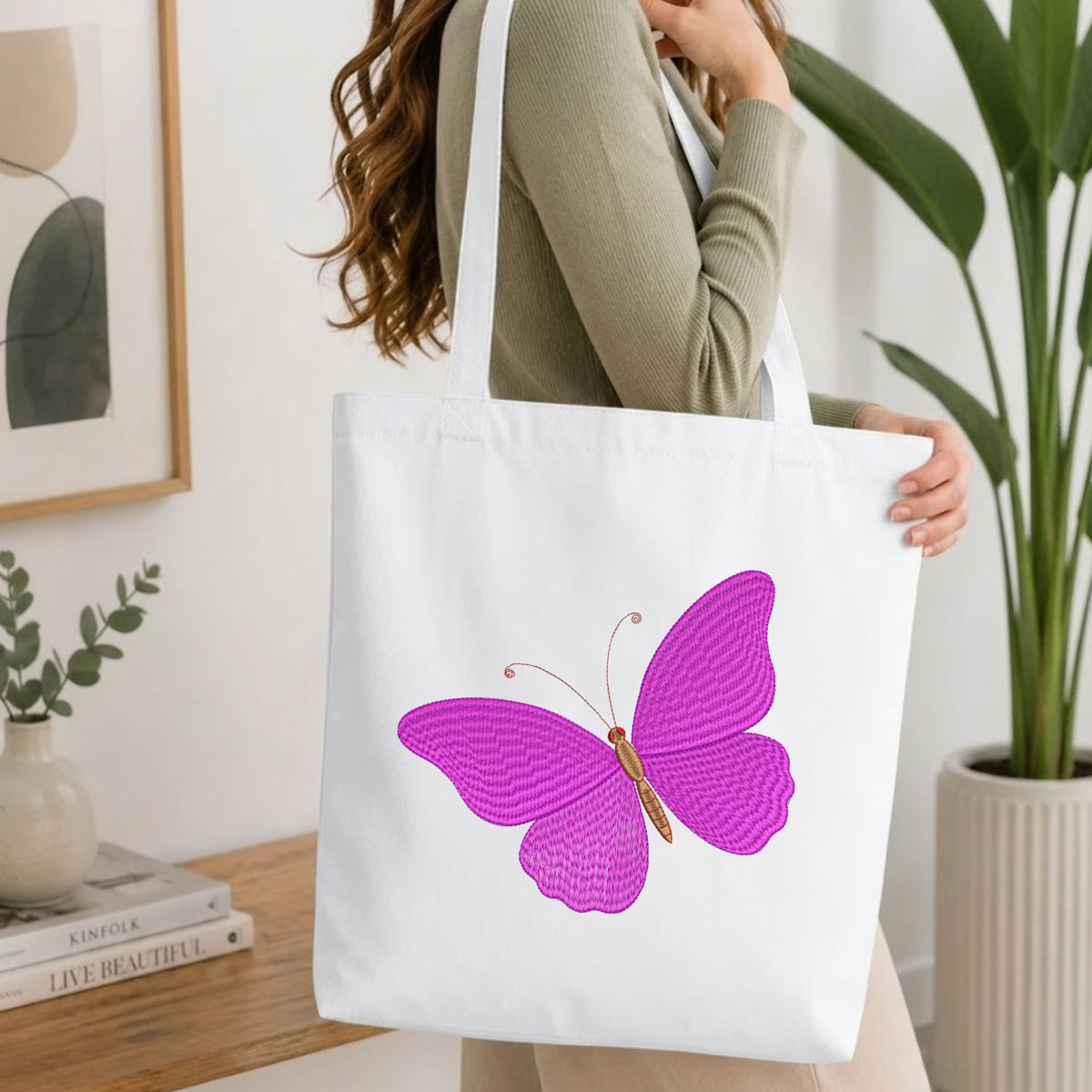 Woman carrying a white natural canvas tote bag featuring an embroidered vibrant purple butterfly design in a bright, modern interior setting.