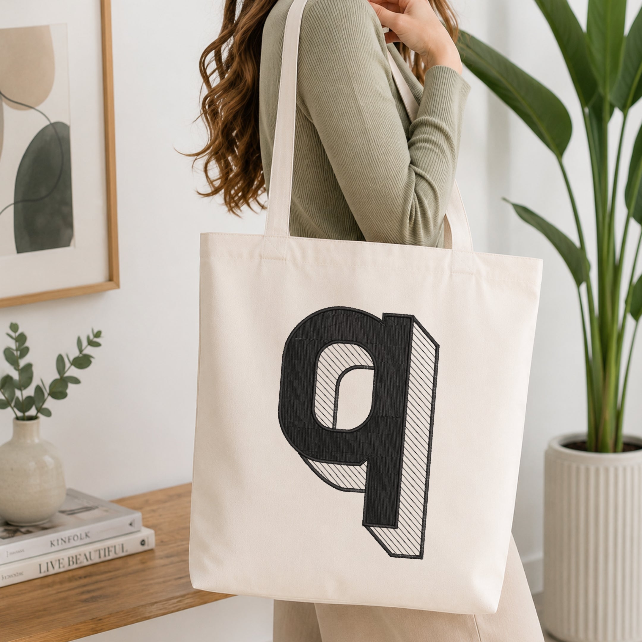 A woman wearing a sage green ribbed long-sleeve top carries a cream canvas tote bag with a large black stylized letter 'q' printed on the front, set in a bright, modern room with a wooden table, framed art, and a potted plant.