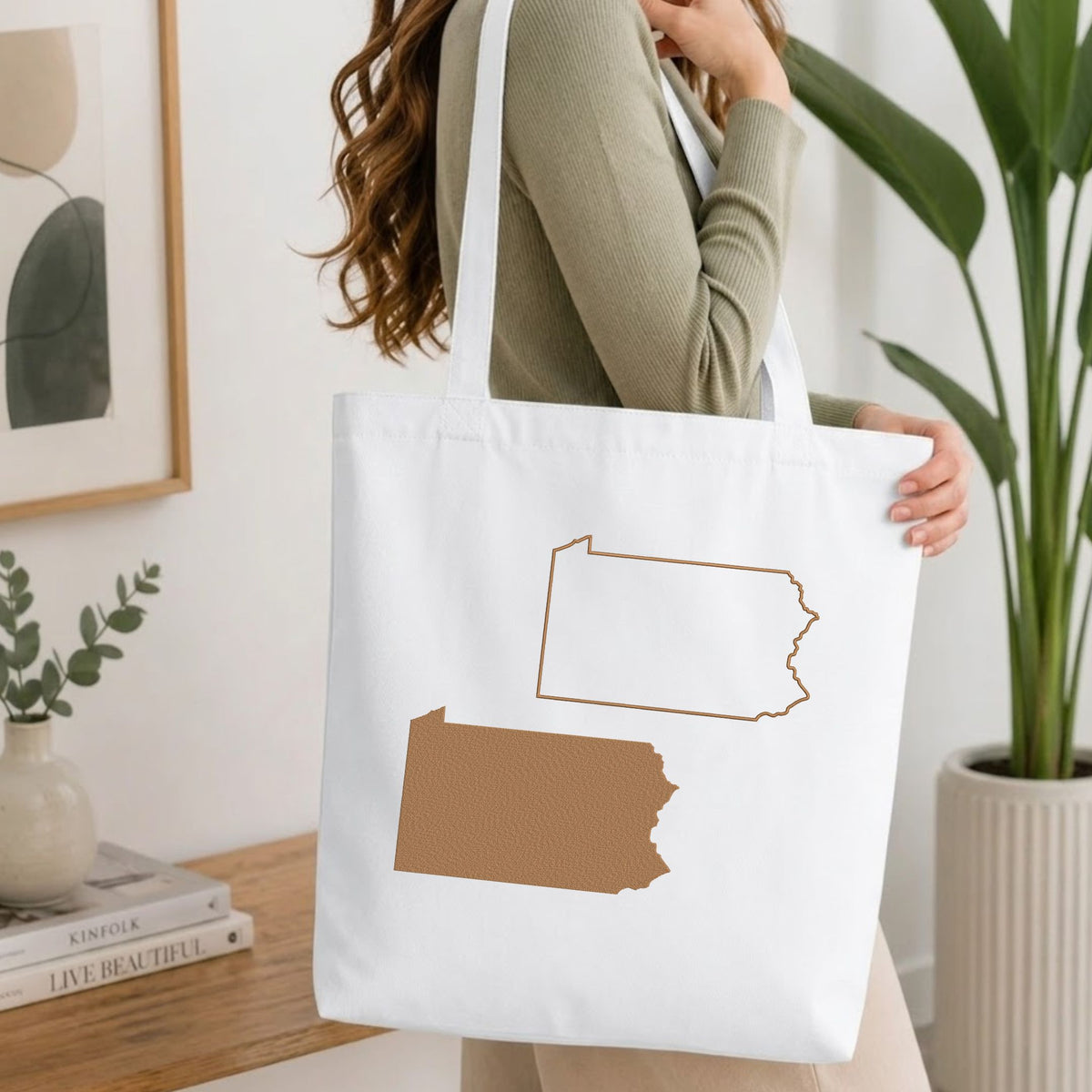 A woman wearing a sage green ribbed long-sleeve top carries a white canvas tote bag with brown outlines of the state of Pennsylvania. The scene is set in a bright, modern room with a wooden table, books, a framed print, and a potted plant.