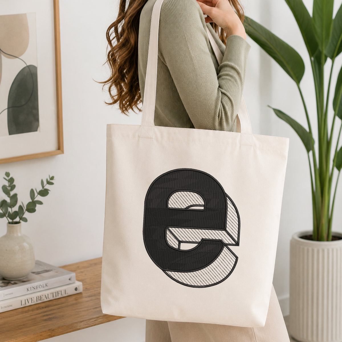 A person holding a natural canvas tote bag printed with an embroidered 3D block lowercase letter "e" design, set against a bright interior room background with green plants.