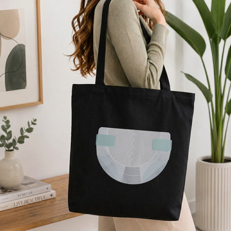 A woman wearing a beige ribbed sweater carries a black canvas tote bag with a large light blue and white diaper graphic on the front, set in a bright, modern room with a potted plant and books.
