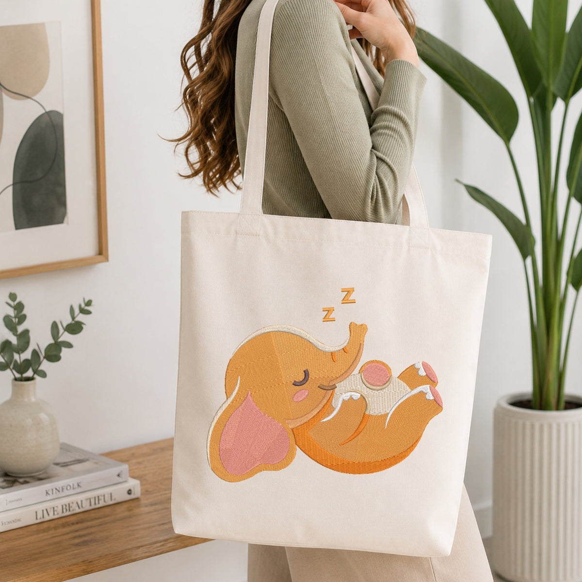 A woman in a sage green ribbed sweater carries a cream canvas tote bag with a large embroidered sleeping orange elephant design on the front, set in a bright, modern room with a wooden shelf and potted plants.