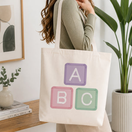 A woman wearing a sage green ribbed sweater carries a cream canvas tote bag with embroidered pastel alphabet blocks reading A, B, and C, in a bright, modern room with a wooden table and potted plants.
