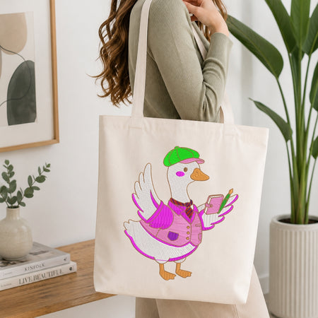 A person carrying a cream-colored tote bag showcasing an embroidered design of a duck wearing a green cap, a pink vest, and holding a small notebook and pen.