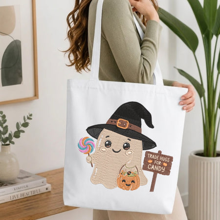 A woman holds a white canvas tote bag featuring a cute cartoon ghost wearing a black witch hat and holding a pumpkin and candy, with a sign that reads 'TRADE HUGS FOR CANDY'. The scene is set in a bright, modern room with a wooden table, books, and a potted plant.