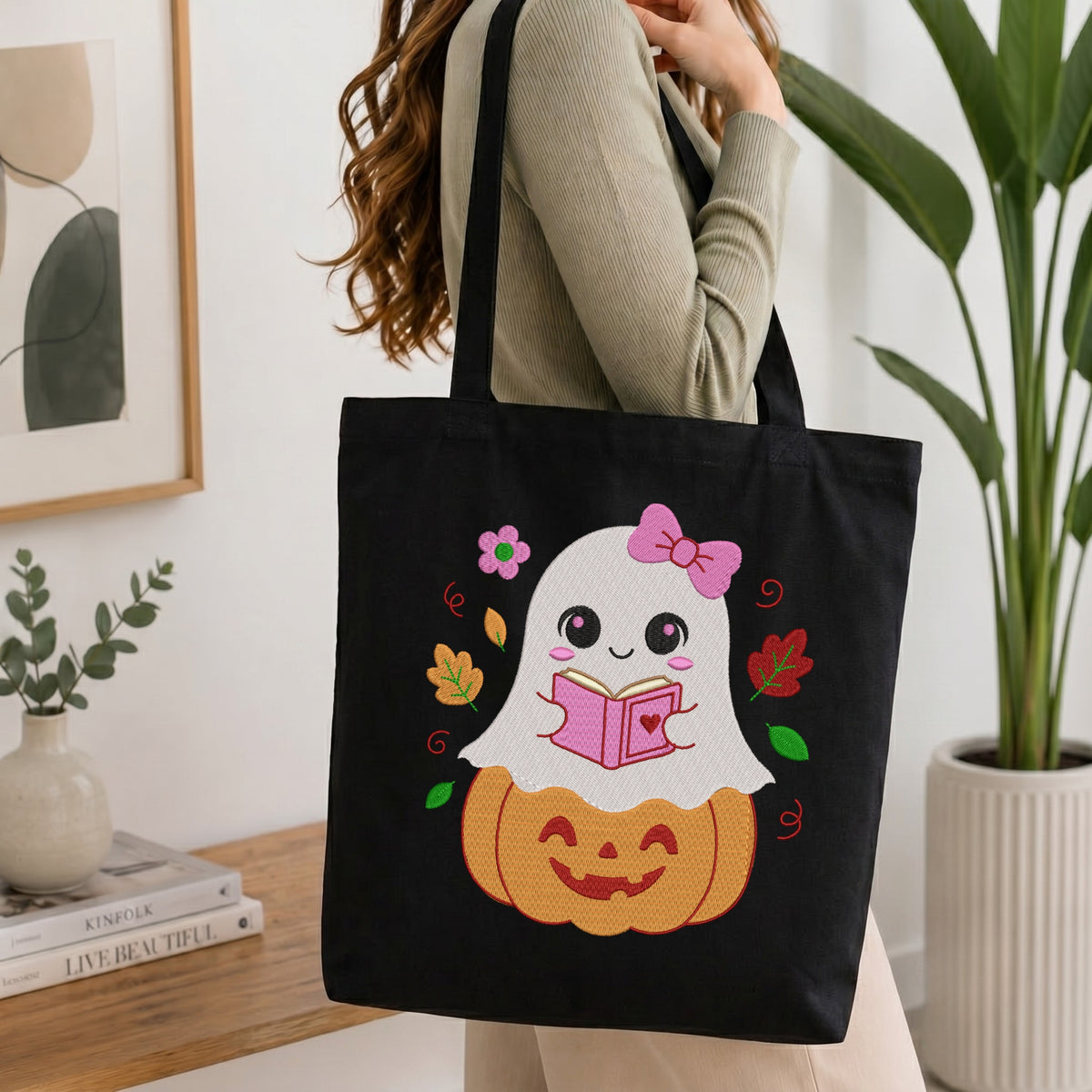 A person holding a black canvas tote bag printed with an embroidered cute reading ghost design, set against a bright interior room background with green plants.